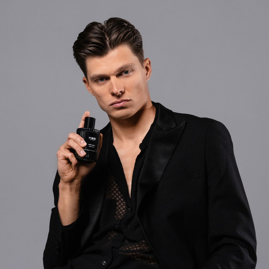 Man in green jacket holding a perfume bottle, seated against a gray background.