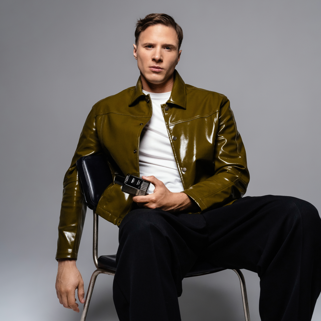 Man in green jacket holding a perfume bottle, seated against a gray background.